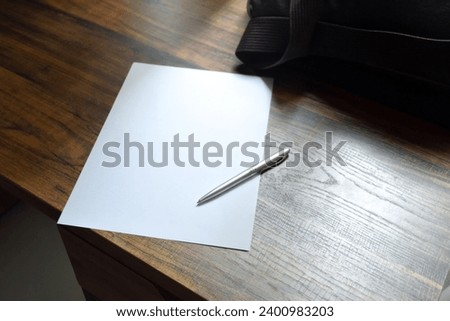 Pen and blank paper and work bag placed on a wooden patterned work desk in soft sunlight background for concept of office and take note.