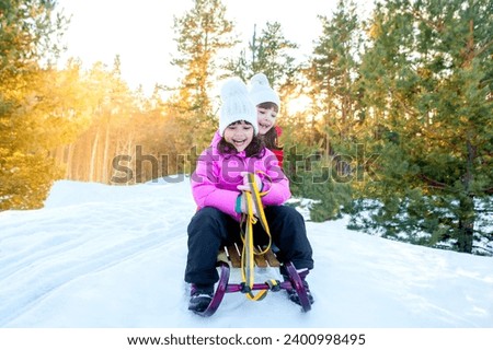Happy funny children ride sleds on snowy road in forest. Family on winter walk. Two little girls are sledding down a hill. Children ride in the snow. Place for text