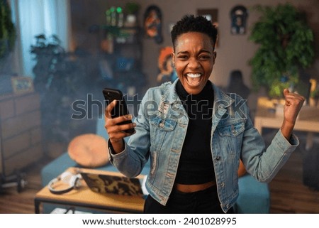 Delighted african american woman celebrating winning lottery holding cell phone in hands. Winner concept. Happy girl doing purchase shopping online payment concept. Royalty-Free Stock Photo #2401028995