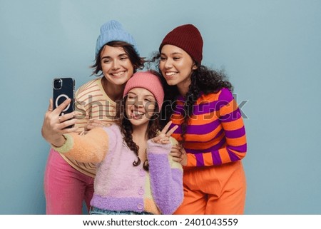 Group of three smiling young girls wearing knit hats taking selfies and gesturing while standing isolated over blue studio background