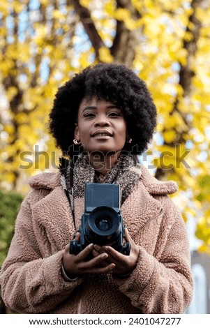 Happy black curly female photographer holding medium format analog camera outdoors. It is autumn or winter cold day and there is a blurry yellow tree behind. She is wearing winter outfit.