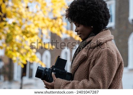 Female curly black photographer using analog film camera in autumn winter day. It is a big medium format camera and she is looking to the waist level finder. Yellow blurry tree on the background.