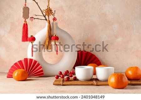Vase with oriental symbols, cups and mandarins on table against beige grunge background. New Year celebration Royalty-Free Stock Photo #2401048149