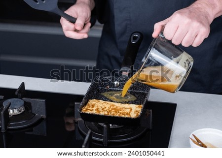 Chef man hands cooking Tamagoyaki or Tamago traditional Japanese Rolled Omelette recipe, made by rolling several layers of cooked scrambled whisking eggs in pan into a rectangular omelet. Royalty-Free Stock Photo #2401050441