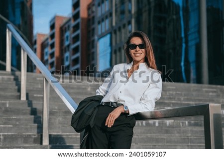 chic businesswoman in a smart suit poses with ease on city steps, her stylish sunglasses reflecting the dynamic architecture around her, showcasing a blend of elegance and corporate life.

 Royalty-Free Stock Photo #2401059107