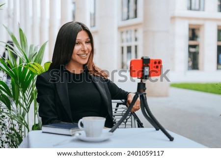 Confident female vlogger with a friendly smile, recording content using a smartphone on a tripod at an outdoor cafe surrounded by nature and modern architecture.