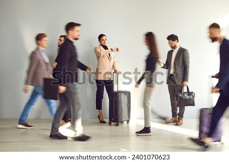 Young woman with a traveling suitcase meeting with a friend, waiting for transport and looking at her watch while standing at the train station or airport among a blurred crowd of walking people