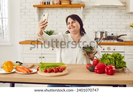 Smiling overweight woman holding bowl of fresh salad and taking selfie. Cheerful plus size woman standing at table in kitchen cooking fresh vegetables for healthy dinner. Healthy eating, diet