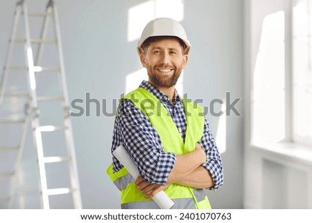 Portrait of smiling builder or architect in hardhat inside new house. Happy bearded man in hard hat and workwear standing with arms folded at home construction site with step ladder in background Royalty-Free Stock Photo #2401070763