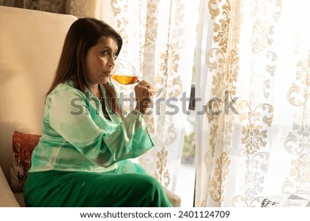 indian woman in casual clothes sitting on wing chair relaxing and drinking wine. lifestyle concept