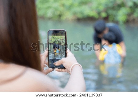 Cropped image of young Asian mom take a picture of a little son and husband playing in river on warm and sunny summer day