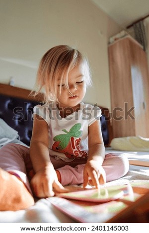 Little girl sits on a bed and reads a book with pictures, running her finger along the lines