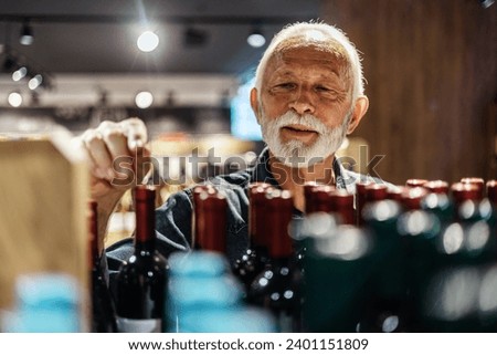 Positive male looking for perfect wine. Man choosing wine bottle picking up from alcohol shelf at wine store shop while he is looking at product and smile.