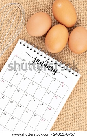 January 2024 Desk calendar and fresh eggs with egg whisk on burlap background.