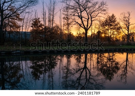 This captivating image depicts a tranquil lake at sunset, where the water acts as a mirror, reflecting the silhouettes of tall, bare trees against a sky painted with the warm, vibrant colors of dusk