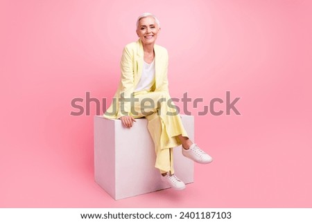 Full size photo of good mood nice cheerful woman dressed yellow suit sit on white cube legs crossed isolated on pink color background