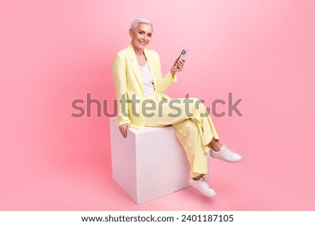 Full size photo of satisfied woman dressed yellow suit sit on white cube read email on smartphone isolated on pink color background