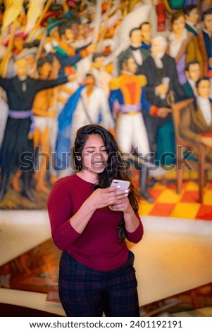 Smiling young Latin woman tourist in casual clothes with dark long hair taking using smartphone in front of mural in Chapultepec Castle in Mexico city