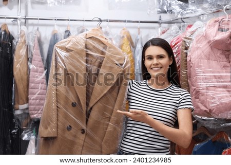Dry-cleaning service. Happy woman holding hanger with coat in plastic bag near racks with clothes indoors Royalty-Free Stock Photo #2401234985