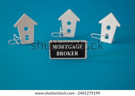 Mortgage broker symbol. Wooden houses sits next to a wooden black board with the word mortgage broker. Beautiful blue background. Business and mortgage broker concept.