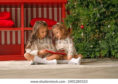 Sisters read New Year's fairy tales sitting on the floor by the Christmas tree. Children look at pictures in a book with stories