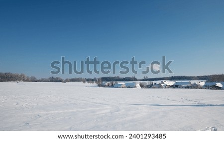 In winter there is snow on the fields. A small village can be seen in the distance. The sky is blue. The sun is shining. There are conifers on the horizon.