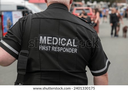 A close-up of a Caucasian male emergency health medical first responder or paramedic wearing a black uniform with grey letters. The ambulance attendant is wearing a short-sleeve shirt with lettering. Royalty-Free Stock Photo #2401330175