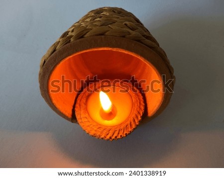 The picture shows an orange wax candle placed in a small brown clay pot. There are notches along the edge around the center with a white string for use in lighting candles in various ceremonies during