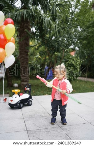 Little girl takes out a toy sword from a scabbard while standing in the garden next to a bunch of balloons Royalty-Free Stock Photo #2401389249