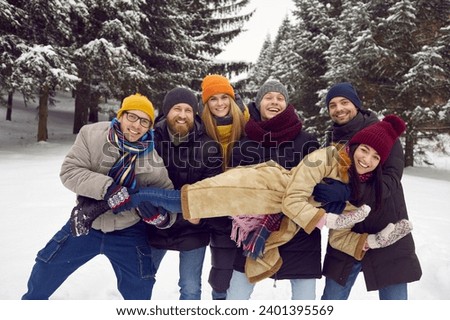 Bunch of happy millennial friends having fun in winter forest. Cheerful young people in warm hats, coats and jackets all together holding one of the girls for funny group photo among snowy fir trees