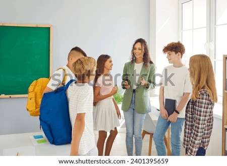 Group of happy school children with their young friendly female teacher standing in a circle in the classroom together and talking after a lesson. Back to school, education and knowledge concept.