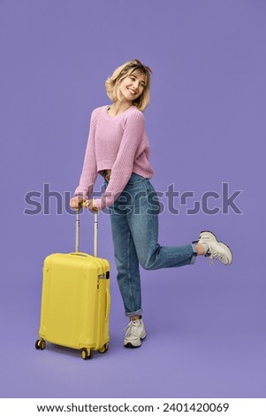 Happy blonde girl tourist holding travel suitcase standing isolated on purple background. Smiling young woman traveler holding baggage ready for summer vacation trip. Full body vertical shot.