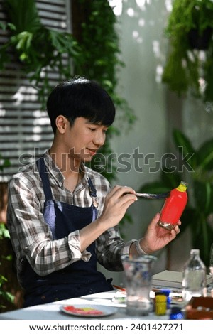 Happy Asian man painting on a glass jar with a brush in workshop. Indoors leisure activity concept