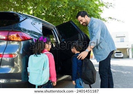 Cheerful father smiling while picking up his young children from elementary school and getting them into the car Royalty-Free Stock Photo #2401451187