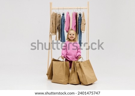 A child, a little girl, stands near the closet, chooses clothes against a light background. Dressing room with clothes on hangers. Wardrobe of children's and stylish clothes. Montessori wardrobe. Royalty-Free Stock Photo #2401475747