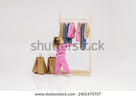 A child, a little girl, stands near the closet, chooses clothes against a light background. Dressing room with clothes on hangers. Wardrobe of children's and stylish clothes. Montessori wardrobe. Royalty-Free Stock Photo #2401475757
