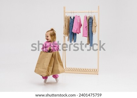 A child, a little girl, stands near the closet, chooses clothes against a light background. Dressing room with clothes on hangers. Wardrobe of children's and stylish clothes. Montessori wardrobe. Royalty-Free Stock Photo #2401475759