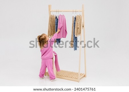 A child, a little girl, stands near the closet, chooses clothes against a light background. Dressing room with clothes on hangers. Wardrobe of children's and stylish clothes. Montessori wardrobe. Royalty-Free Stock Photo #2401475761