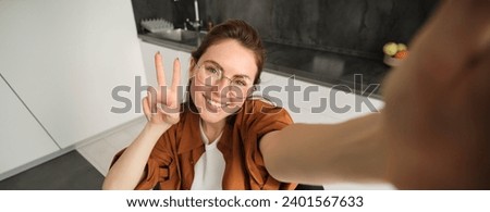 Portrait of stylish young woman at home, taking selfie on smartphone, posing in kitchen and smiling at mobile camera.