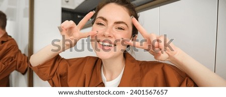 Close up portrait of happy, beautiful girl listens to music in wireless headphones, shows peace, v-sign gestures and smiles, enjoys songs in favourite playlist.