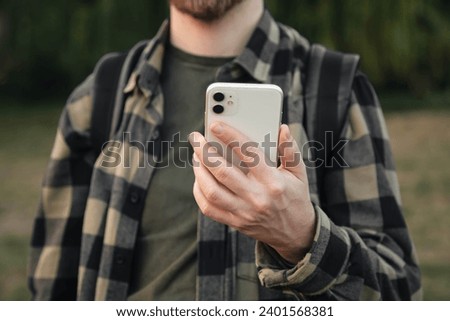 A man in a green shirt uses a white smartphone on a blurred background of nature outside, technology use concept, copy space.