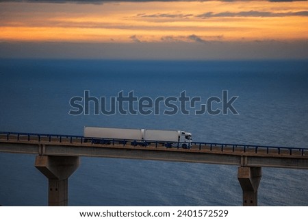 Mega-trailer truck driving over a bridge with the sea and dawn sky on the horizon. Royalty-Free Stock Photo #2401572529