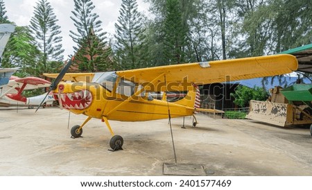 Vehicle photos Small yellow classic fancy airplane The plane's head is shaped like a sharp dinosaur tooth. Parked on a wide, cement floor. Behind there are many tall pine trees. During the day the sky