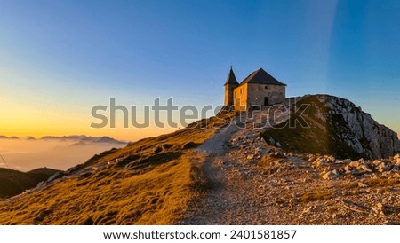 Scenic sunrise view of chapel Maria am Stein on top of mountain peak Dobratsch, Villacher Alps, Austria, Europe. Looking at Julian and Karawanks mountain range. Golden morning hour tranquil atmosphere Royalty-Free Stock Photo #2401581857