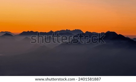 Panoramic sunrise view from Dobratsch on Julian Alps and Karawanks in Austria, Europe. Silhouette of endless mountain ranges with orange and pink sky. Jagged sharp peaks and valleys. Cottage on hill Royalty-Free Stock Photo #2401581887
