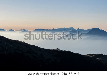 Panoramic sunrise view from Dobratsch on Julian Alps and Karawanks in Austria, Europe. Silhouette of endless mountain ranges with orange and pink sky. Jagged sharp peaks and valleys. Cottage on hill Royalty-Free Stock Photo #2401581917