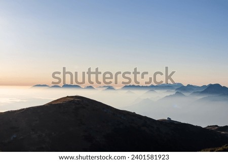 Panoramic sunrise view from Dobratsch on Julian Alps and Karawanks in Austria, Europe. Silhouette of endless mountain ranges with orange and pink sky. Jagged sharp peaks and valleys. Cottage on hill Royalty-Free Stock Photo #2401581923