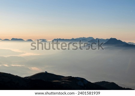 Panoramic sunrise view from Dobratsch on Julian Alps and Karawanks in Austria, Europe. Silhouette of endless mountain ranges with orange and pink sky. Jagged sharp peaks and valleys. Cottage on hill Royalty-Free Stock Photo #2401581939