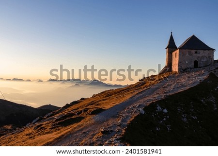 Scenic sunrise view of chapel Maria am Stein on top of mountain peak Dobratsch, Villacher Alps, Austria, Europe. Looking at Julian and Karawanks mountain range. Golden morning hour tranquil atmosphere Royalty-Free Stock Photo #2401581941