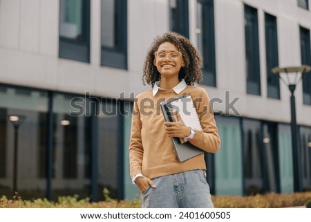 Smiling female freelancer standing with laptop and note pads on modern building backgrounds